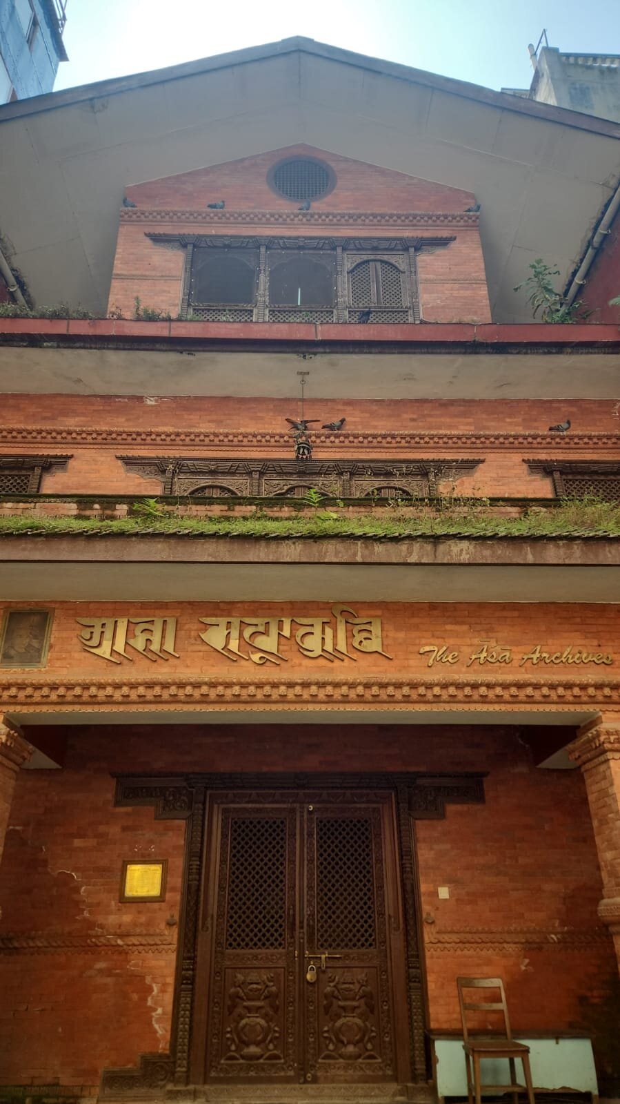 The brick façade of the Āśā Saphū Kuthi, with carved wooden windows and the library's name in gilt Nepāl-script letters over the porch